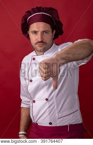 Unshaven Handsome Male Chef Cook Shows Thumb Down Looking At Camera Isolated On Red Backdrop. Chef D