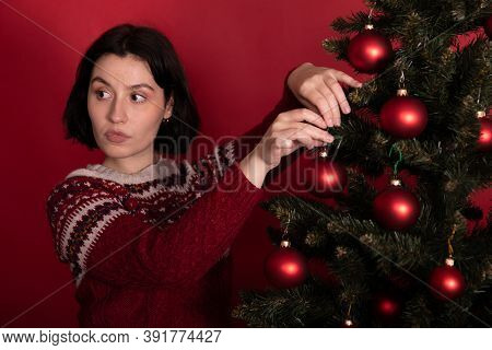 Thoughtful Brunette Girl Dressed In Knitted Winter Pattern Sweater Hanging Toy Balls On Christmas Tr