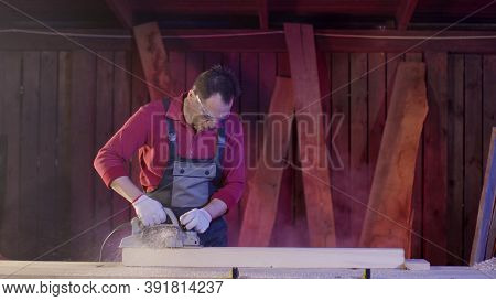 Man Carpenter Cleans The Surface Of Wooden Beam Using Electric Planer In Workshop And Flies Sawdust.