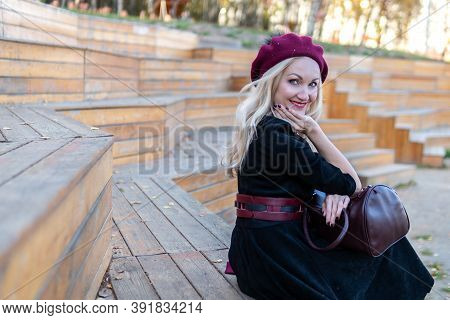 Happy Sits On A Bench In A Summer Theater, Wooden In A Burgundy Coat And Biret, With Tender Lips, In