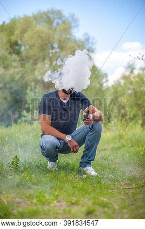 Bearded Man Blowing A Lot Of Smoke Using Vape Electronic Cigarette On The Forest Ground. Vaping Acti