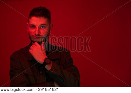 Low Key Portrait Of Bearded Young Man Looking At Camera Ironic Expression Dressed Casually In Red Li