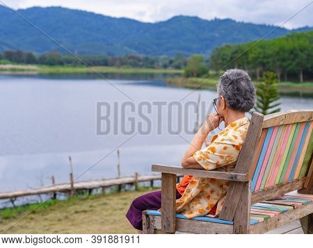 Side View Of A Senior Woman While Sitting On Chairside The Lake. Space For Text
