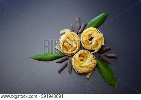 Tagliatelle Pasta, Brown Penne Rigate Pasta And Laurel Leaves On A Gray Background.