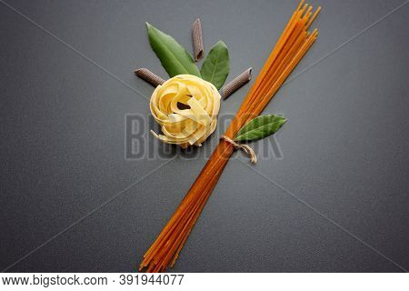 Tagliatelle Pasta, Brown Penne Rigate Pasta,  Brown Spaghetti And Laurel Leaves On A Gray Background