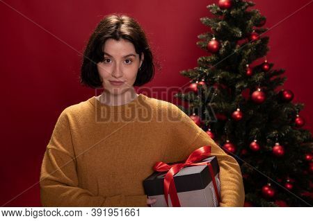 Portrait Of Young Pretty Woman Holding Xmas Gift Box Looks With Smile At Camera On Red Background Wi