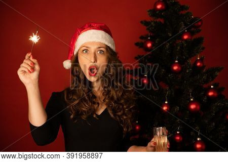 Portrait Of Pretty Surprised Woman In Santa Hat Holding Burning Sparkler On Xmas Tree Red Background