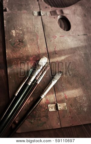 Old painting brushes and a wooden palette on an old painters desk. 