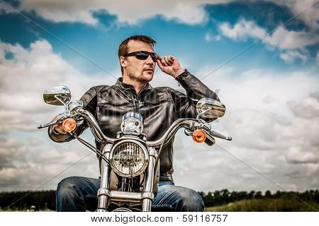 Biker man wearing a leather jacket and sunglasses sitting on his motorcycle looking at the sunset.