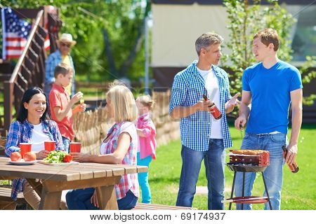 Portrait of young men frying sausages and talking in the countryside at weekend with their families near by
