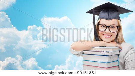education, high school, knowledge, and people concept - picture of happy student girl or woman in trencher cap with stack of books over blue sky and clouds background