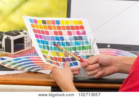 Male hands holding up palette, colormap spread out in front of laptop on working desk