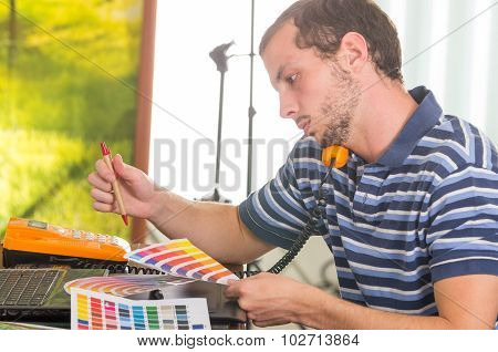 Man wearing blue white striped t-shirt sitting by work desk using phone and looking at palette, colo