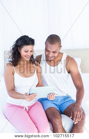Happy husband with pregnant wife holding digital tablet while sitting on bed at home