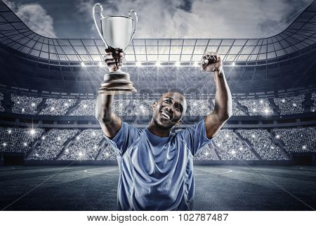 Happy sportsman looking up and cheering while holding trophy against large football stadium under spotlights
