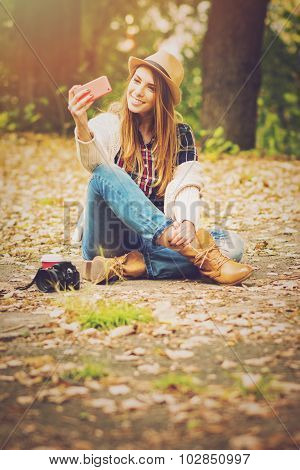 Beautiful fashionable teenage girl taking a selfie with smartphone sitting in park in autumn with di