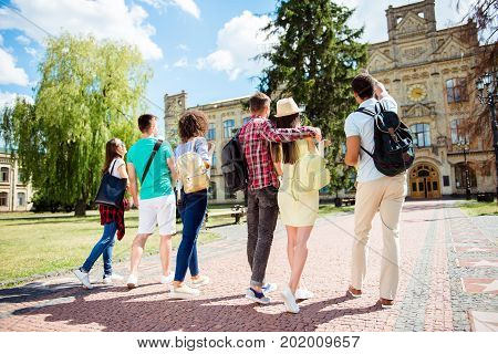 Rear Low Angle View Of Six Multiethnic Students. They Are Walking To College Building And Discuss Th