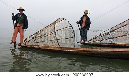 Inle Lake Myanmar - January 04 2007: Two local fishers with traditional Burmese fishing nets standing on stern of long boats and rowing oars by leg on Inle lake Shan state Myanmar.