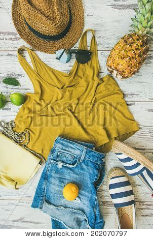Colorful summer fashion outfit flat-lay. Denim shorts, straw sun hat, yellow top, espadrillas, yellow leather bag, sunglasses, pineapple, limes and lemon over pastel parquet background, top view