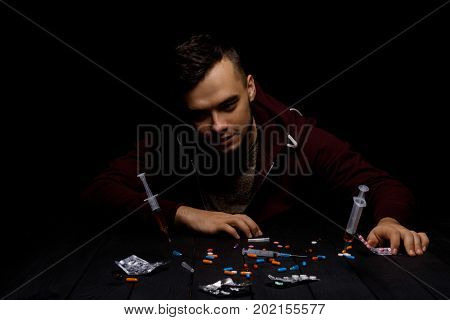 An addict male clings to his head with his own hands sitting at the table on a black background. A hopeless drug addict is going through addiction crisis. A lot of colorful narcotics on a desk.