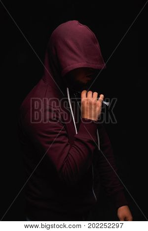 A photo of a desperate young drug addict is wearing a hood on a saturated black background. A hopeless addict male with a syringe in his hand. An addict injects a narcotic substance into the neck.