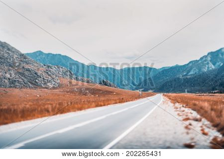 Tilt-shift shooting of road stretching between dry autumn meadows and mountain ridges overgrown with trees power line on the right Altai mountains near Kuyus district Russia
