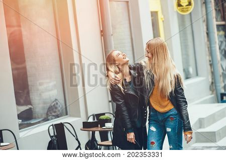 It is funny walk with best friend! Two beautiful women walking outdoor hugging and laughing on autumn street. Fashion urban girls spending weekend talking at sunset near a cafe.