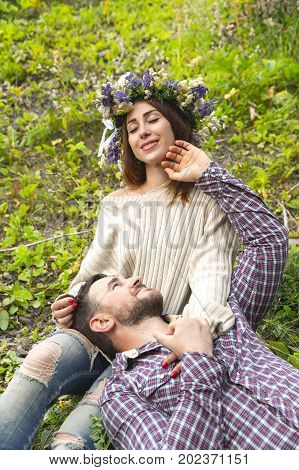 Hipster with a beard lies on the lap of his girl who wears a vinok of wildflowers. The concept of unity with nature and a young couple of people