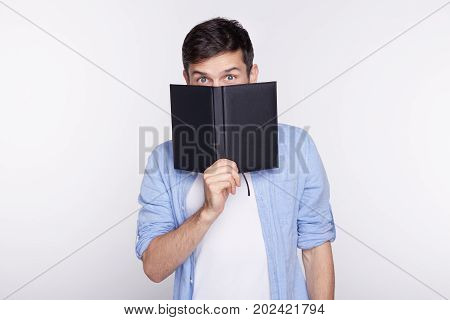 Studio shot of European blue eyed businessman or student dressed casual hiding face behind notebook while standing isolated on white backgroundlooking humorously at camera. People business career.