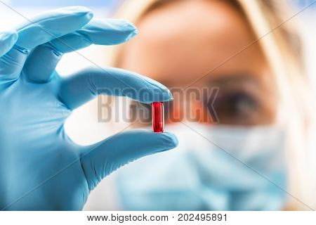 Young attractive female scientist with protective eyeglasses and mask holding a red transparent pill with fingers in gloves in the pharmaceutical research laboratory