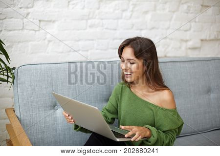 Attractive young Latin woman dressed in casual wear enjoying online communication sitting on grey couch at home and having video conference call with friends using web enabled laptop computer