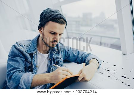 Serious young student is sitting on sofa and writing down some plas. He is making his notes