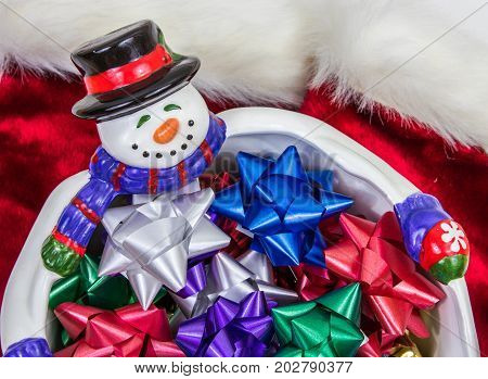A top down horizontal photo of a smiling snowman bowl with lots of colorful bows in it and a red and white velvet background