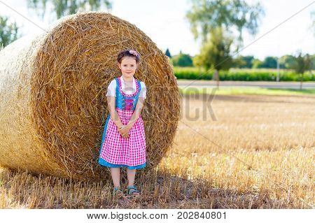 Cute little kid girl in traditional Bavarian costume in wheat field. German child with hay bale during Oktoberfest in Munich. Preschool girl play at hay bales during summer harvest time in Germany