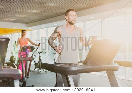 Young happy handsome man with tattoo in fitness club. Cardio workout, running on treadmill. Healthy lifestyle, guy training in gym. Flare effect