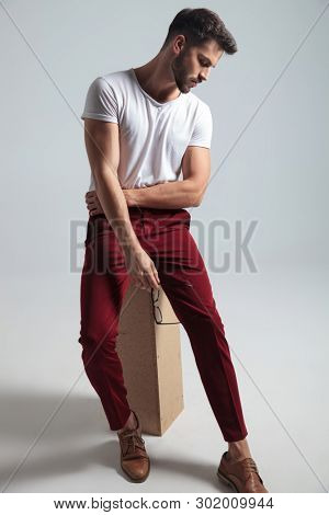 Concerned man holding his glasses and looking downwards while wearing a white t-shirt and red pants, sitting on gray studio background