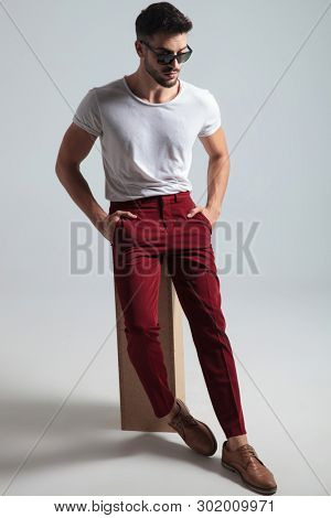 Thoughtful boy looking down and holding both of his hands in his pockets while wearing a white t-shirt, red pants and sunglasses, sitting on gray studio background