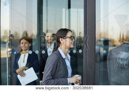 Smiling Attractive Young Businesswoman In Glasses Holding Coffee Cup And Walking Through Revolving D