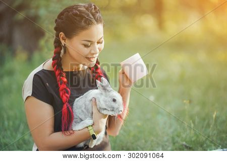 Pretty Asian Girl Drinks Tea And Huggs Rabbit On Green Grass Of Summer Forest. Good-looking Girl Wit