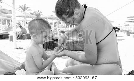 Black And White Image Of Young Caring Mother Applying Sunscreen Lotion On Her Little Child On The Be
