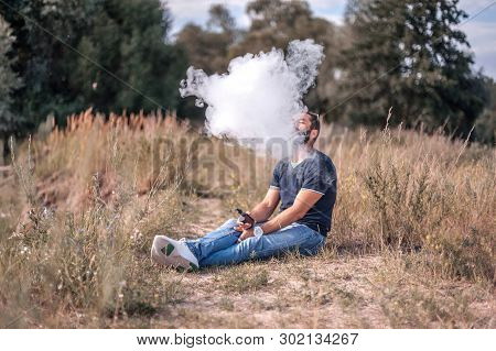 Bearded Smoker Blowing A Lot Of Smoke Using Vape Electronic Cigarette In Nature. Electronic Cigarett