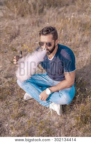 Stylish Bearded Vaper Enjoying An Electronic Cigarette In The Woods. The Man Smokes.