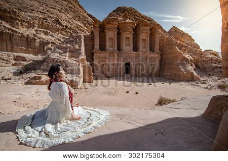 Asian Woman Tourist In White Dress Sitting At Ad Deir Or El Deir, The Monument Carved Out Of Rock In