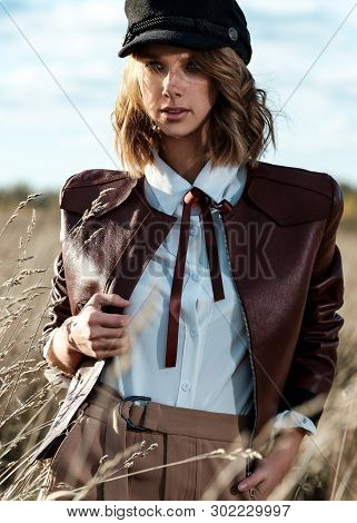 Portrait Of Attractive Female Model In Leather Jacket And Fashionable Cap Looking At Camera Against 