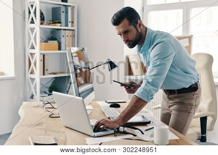 Inspired To Work Hard. Young Modern Businessman Using Smart Phone And Computer While Working In The 