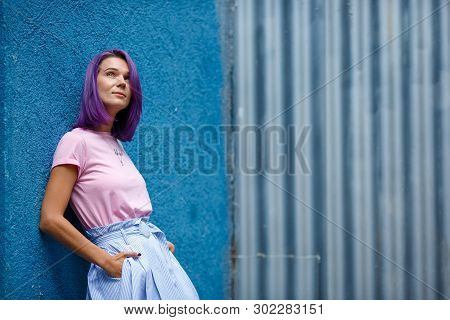 Slim Beautiful Girl With Purple Hair On A Blue Background, Is Leaning On The Wall Of The Building, L