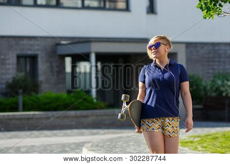 Stylish Girl Plus Size In Blue T-shirt And Shorts And Sunglasses With A Longboard In Her Hands Goes 
