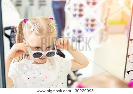 Cute Little Young Caucasian Blond Girl Trying On And Choosing Sunglasses In Front Of Mirror At Optic