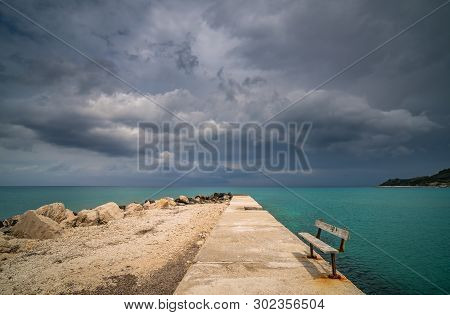 Empty Wooden Bench On A Concrete Pier With Stormy Dramatic Clouds Accumulating Over Alykes Bay In Za