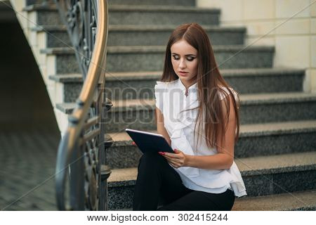 Young And Attractive Female Sittind On The Stairs And Use Tablet. Woman Play Online Games On Tablet